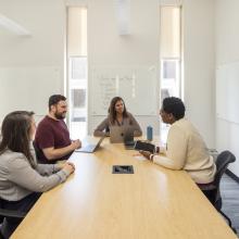 A diverse group of four Penn GSE students and staff sit around a board table with laptops and tablets talking. A whiteboard lists topics on the wall behind them.